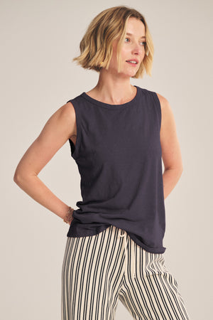 Woman with short blonde hair wears the TAURUS TANK TOP by Velvet by Graham & Spencer, paired with white pants featuring black vertical stripes, standing against a neutral background.