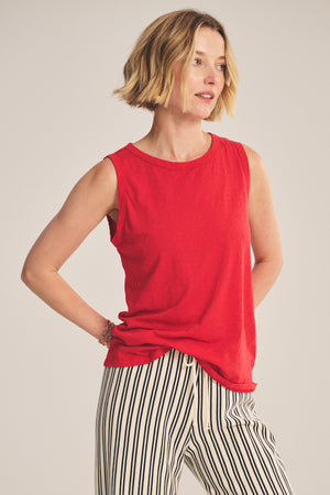 A woman wears the TAURUS TANK TOP by Velvet by Graham & Spencer—a sleeveless red crew neck—paired with black-and-white vertically striped pants, standing against a plain background.