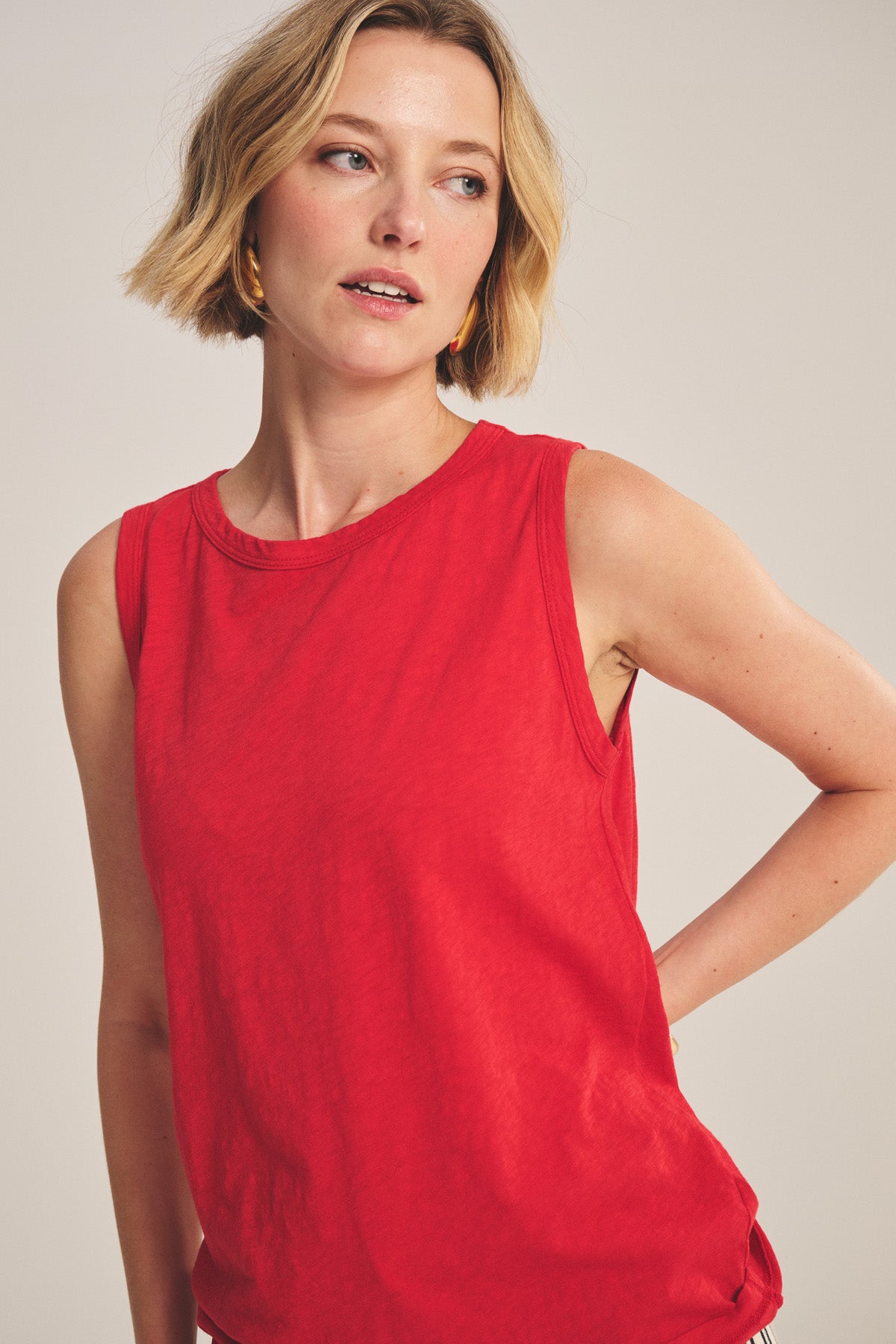 A woman with short blonde hair models the TAURUS TANK TOP by Velvet by Graham & Spencer, a sleeveless red essential, standing against a plain light background and glancing to the side.