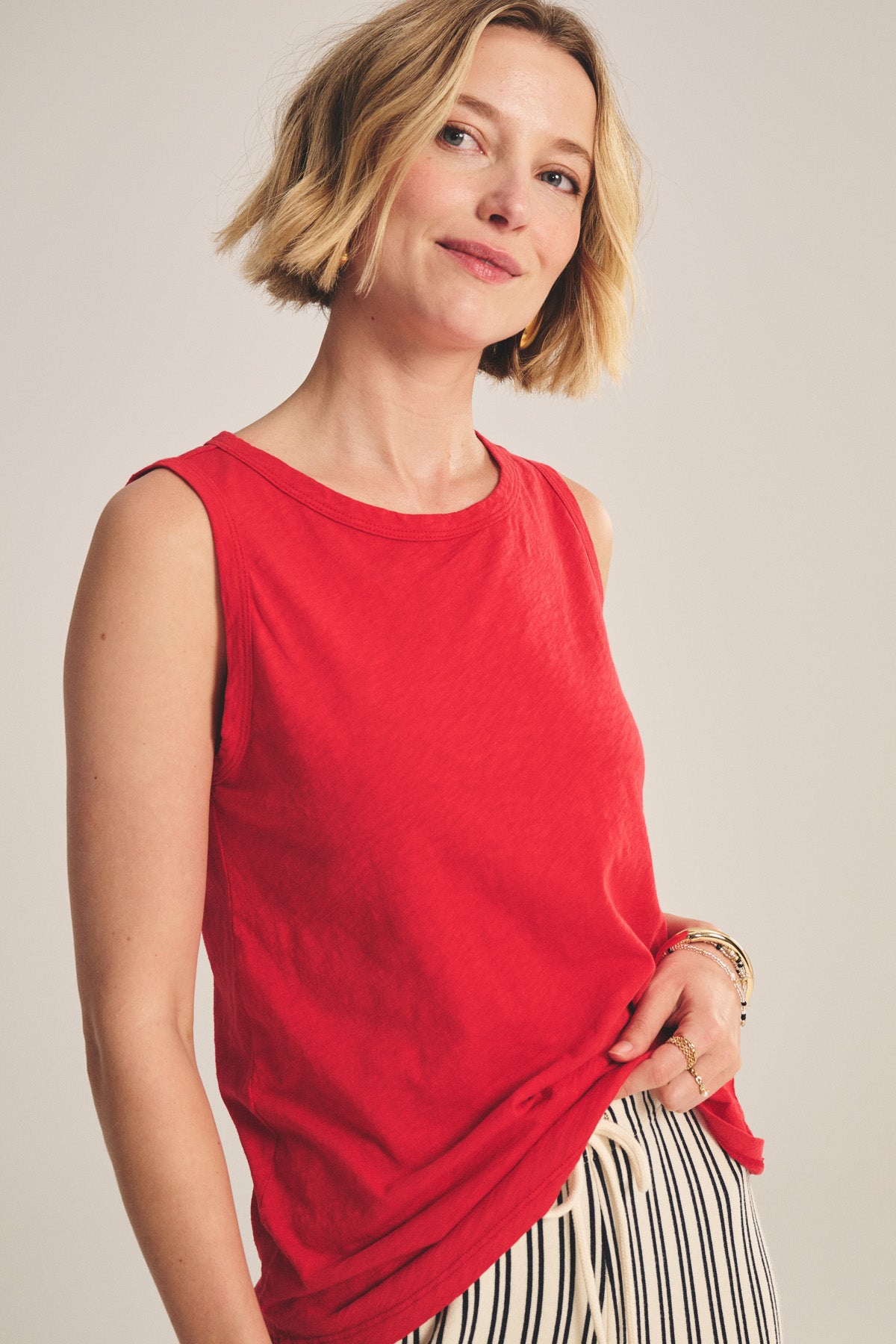 A woman with a blonde bob wears the TAURUS TANK TOP from Velvet by Graham & Spencer, a sleeveless red cotton slub style, paired with black-and-white striped pants against a neutral background.-41089089634497