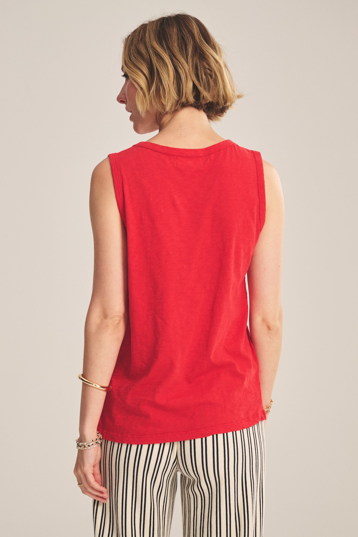 A woman with short blonde hair, seen from behind, wears the TAURUS TANK TOP by Velvet by Graham & Spencer—a sleeveless red crew neck style—paired with black-and-white striped pants.