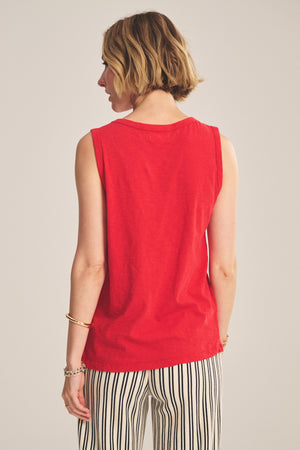 A woman with short blonde hair, seen from behind, wears the TAURUS TANK TOP by Velvet by Graham & Spencer—a sleeveless red crew neck style—paired with black-and-white striped pants.