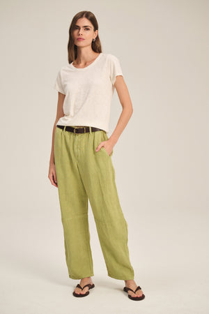 A woman stands against a plain background wearing a white t-shirt, brown sandals, and Velvet by Graham & Spencer’s TAYLOR HEAVY LINEN PANT in light green with a black belt.