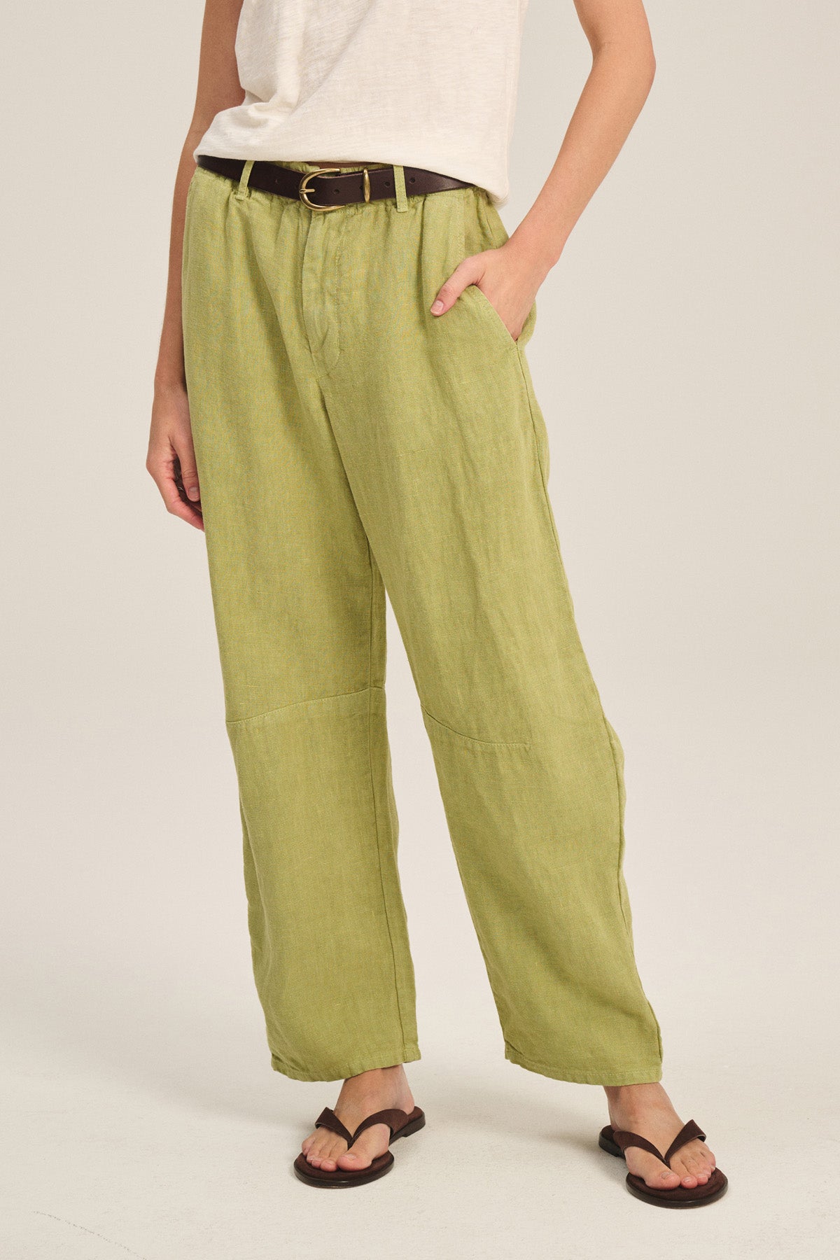 Someone wearing Velvet by Graham & Spencer's TAYLOR HEAVY LINEN PANT in light green, paired with a white tee, dark belt, and black sandals, stands with one hand in their pocket.-41436030632129