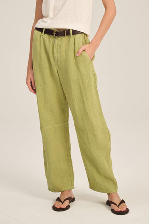 Someone wearing Velvet by Graham & Spencer's TAYLOR HEAVY LINEN PANT in light green, paired with a white tee, dark belt, and black sandals, stands with one hand in their pocket.