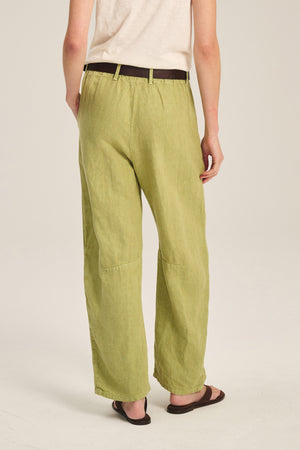 A person, back to camera, wears a beige short-sleeve shirt, dark belt, brown sandals, and the TAYLOR HEAVY LINEN PANT by Velvet by Graham & Spencer in light green with a tailored barrel leg fit.