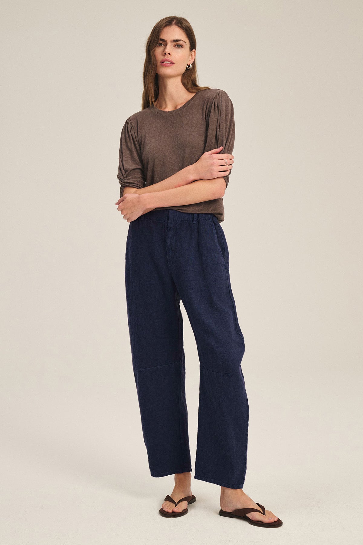 A person stands with arms crossed, wearing a brown T-shirt, brown sandals, and the TAYLOR HEAVY LINEN PANT by Velvet by Graham & Spencer in dark blue with a barrel leg silhouette against a plain light background.