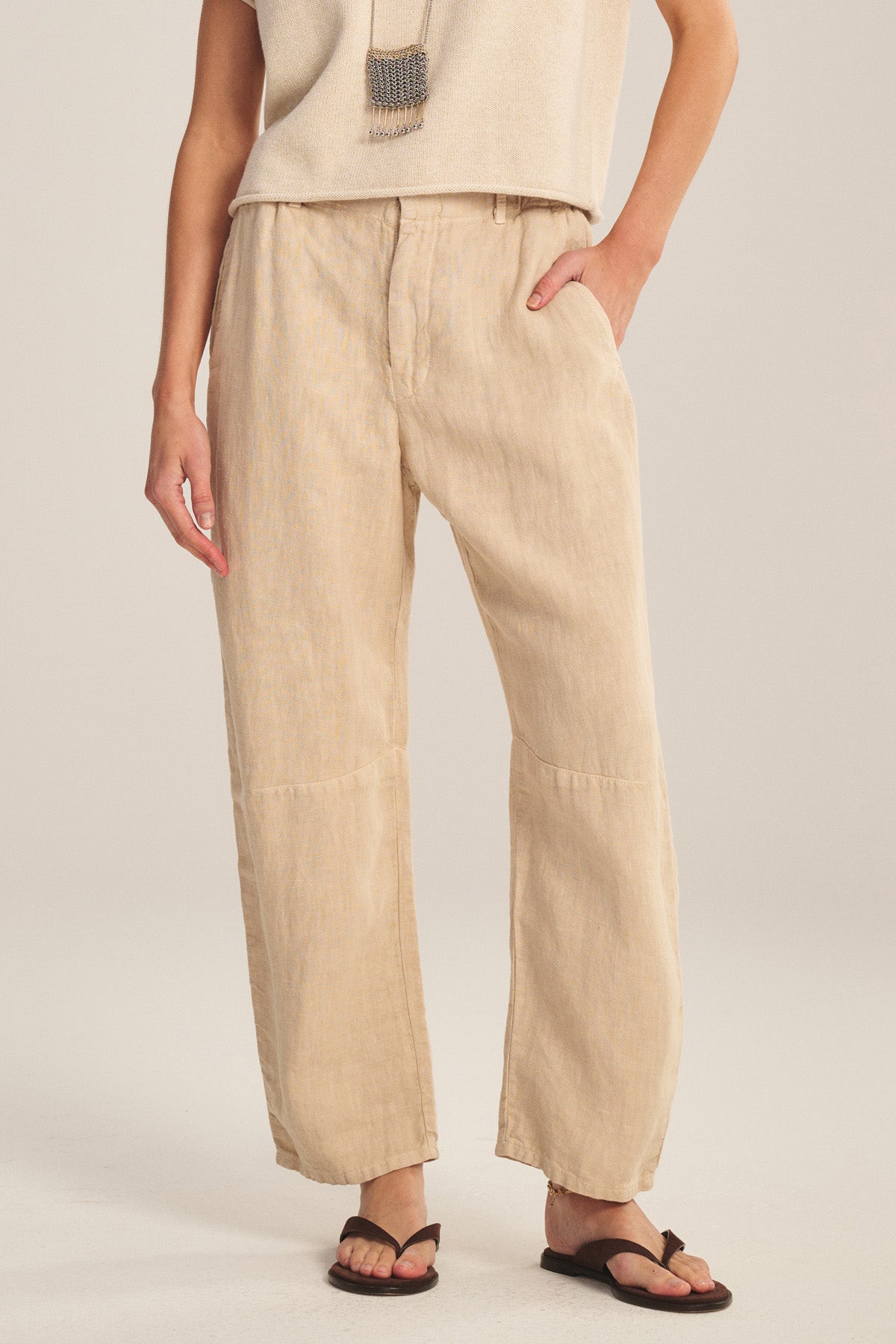 A person wearing Velvet by Graham & Spencer's TAYLOR HEAVY LINEN PANT in tailored light beige with a curved leg, paired with a cream sleeveless top, black sandals, and a long beaded necklace, stands against a neutral background.