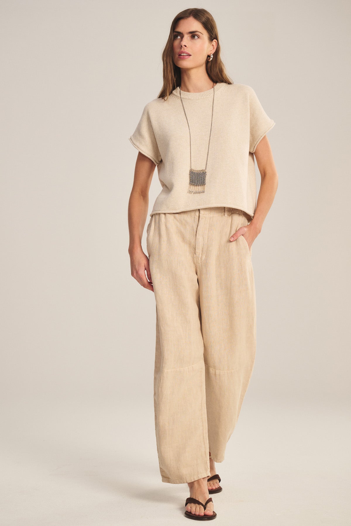 A woman stands against a plain background wearing the TAYLOR HEAVY LINEN PANT by Velvet by Graham & Spencer in beige, paired with a beige short-sleeve top, black sandals, and a long necklace.