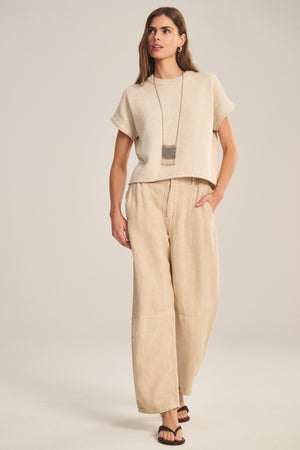 A woman stands against a plain background wearing the TAYLOR HEAVY LINEN PANT by Velvet by Graham & Spencer in beige, paired with a beige short-sleeve top, black sandals, and a long necklace.