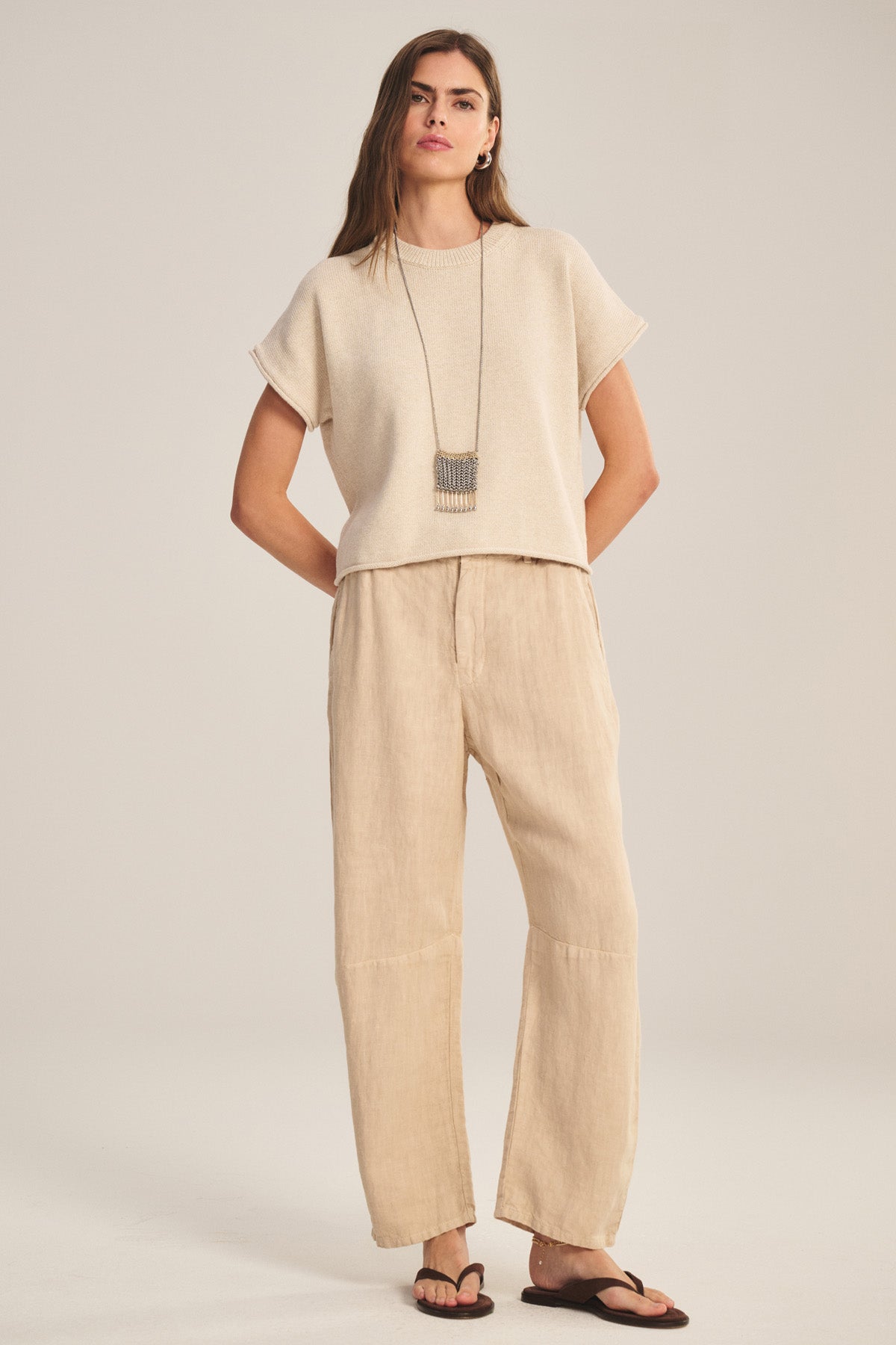 A woman wears the TAYLOR HEAVY LINEN PANT by Velvet by Graham & Spencer, paired with a beige short-sleeve top, black sandals, and a long necklace, standing against a plain light background.