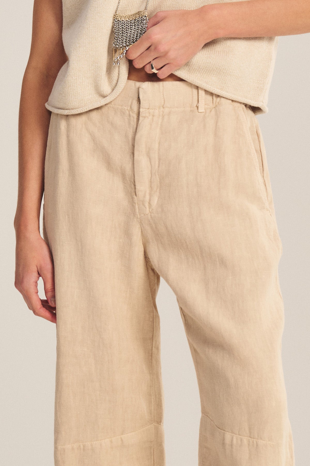 A person wearing the TAYLOR HEAVY LINEN PANT by Velvet by Graham & Spencer in beige, featuring a curved leg silhouette, paired with a light-colored sleeveless top and one hand resting near the waistband.