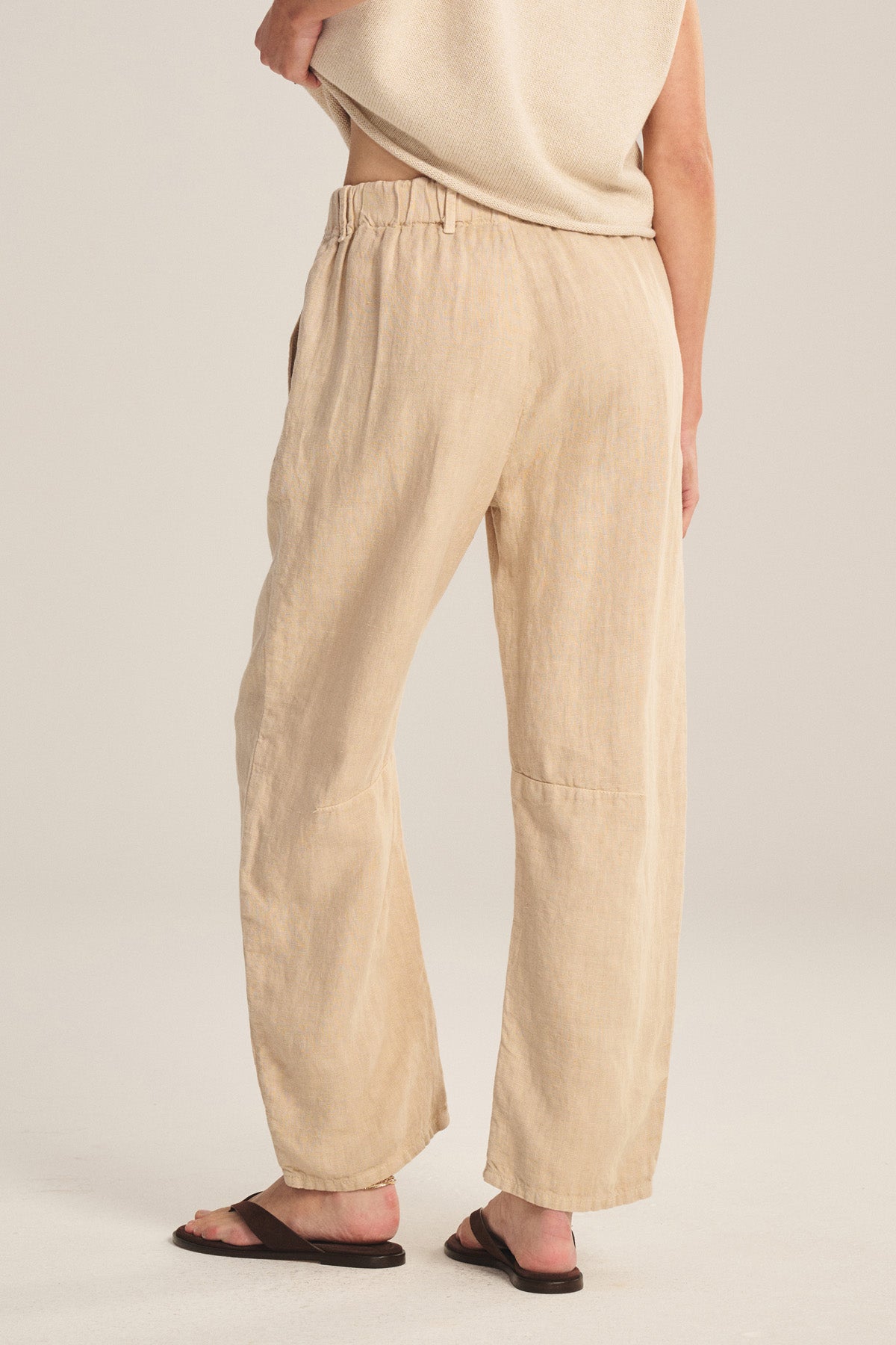 A person is shown from the back wearing the TAYLOR HEAVY LINEN PANT by Velvet by Graham & Spencer—loose, beige pants with a curved leg silhouette—paired with black sandals and a glimpse of a sleeveless top.