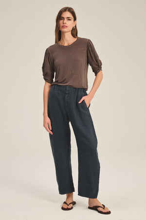 A woman stands against a neutral background wearing a brown t-shirt, flip-flops, and the TAYLOR HEAVY LINEN PANT by Velvet by Graham & Spencer, with one hand in her pocket.
