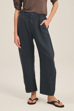 A person wearing Velvet by Graham & Spencer’s TAYLOR HEAVY LINEN PANT in dark blue, styled with a brown long-sleeve top and sandals, stands with one hand in their pocket against a plain background.