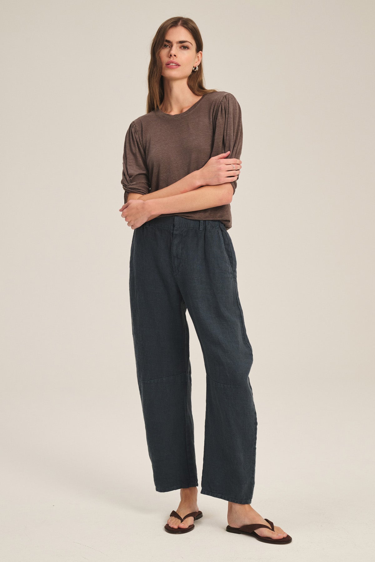 A person stands against a plain background wearing a brown top, Velvet by Graham & Spencer's TAYLOR HEAVY LINEN PANT in a tailored barrel leg fit, and brown sandals, with arms crossed and a neutral expression.