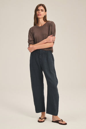 A person stands against a plain background wearing a brown top, Velvet by Graham & Spencer's TAYLOR HEAVY LINEN PANT in a tailored barrel leg fit, and brown sandals, with arms crossed and a neutral expression.