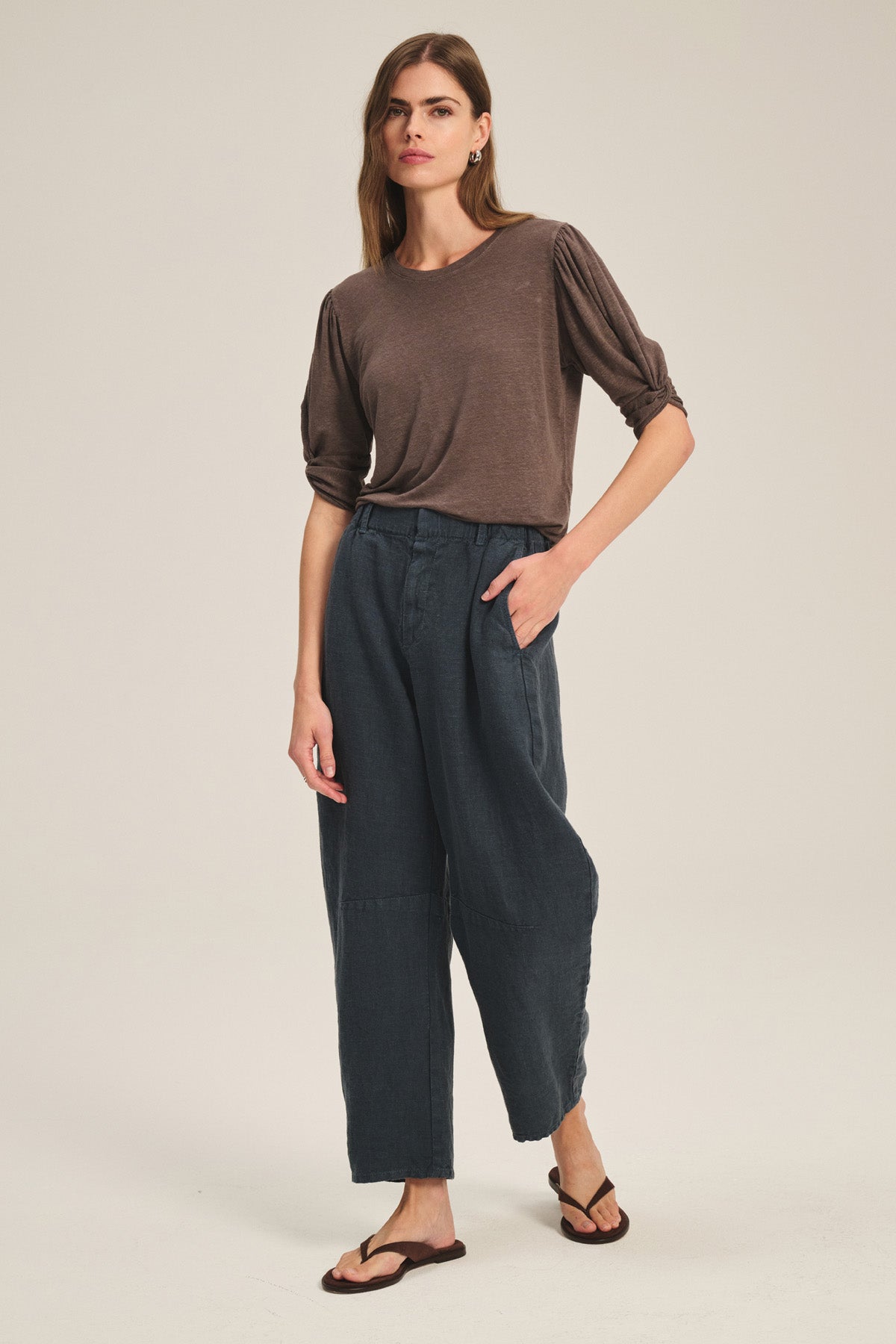A woman stands against a plain background wearing a loose brown top, brown sandals, and the TAYLOR HEAVY LINEN PANT by Velvet by Graham & Spencer, with one hand in her pocket.