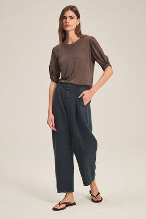 A woman stands against a plain background wearing a loose brown top, brown sandals, and the TAYLOR HEAVY LINEN PANT by Velvet by Graham & Spencer, with one hand in her pocket.
