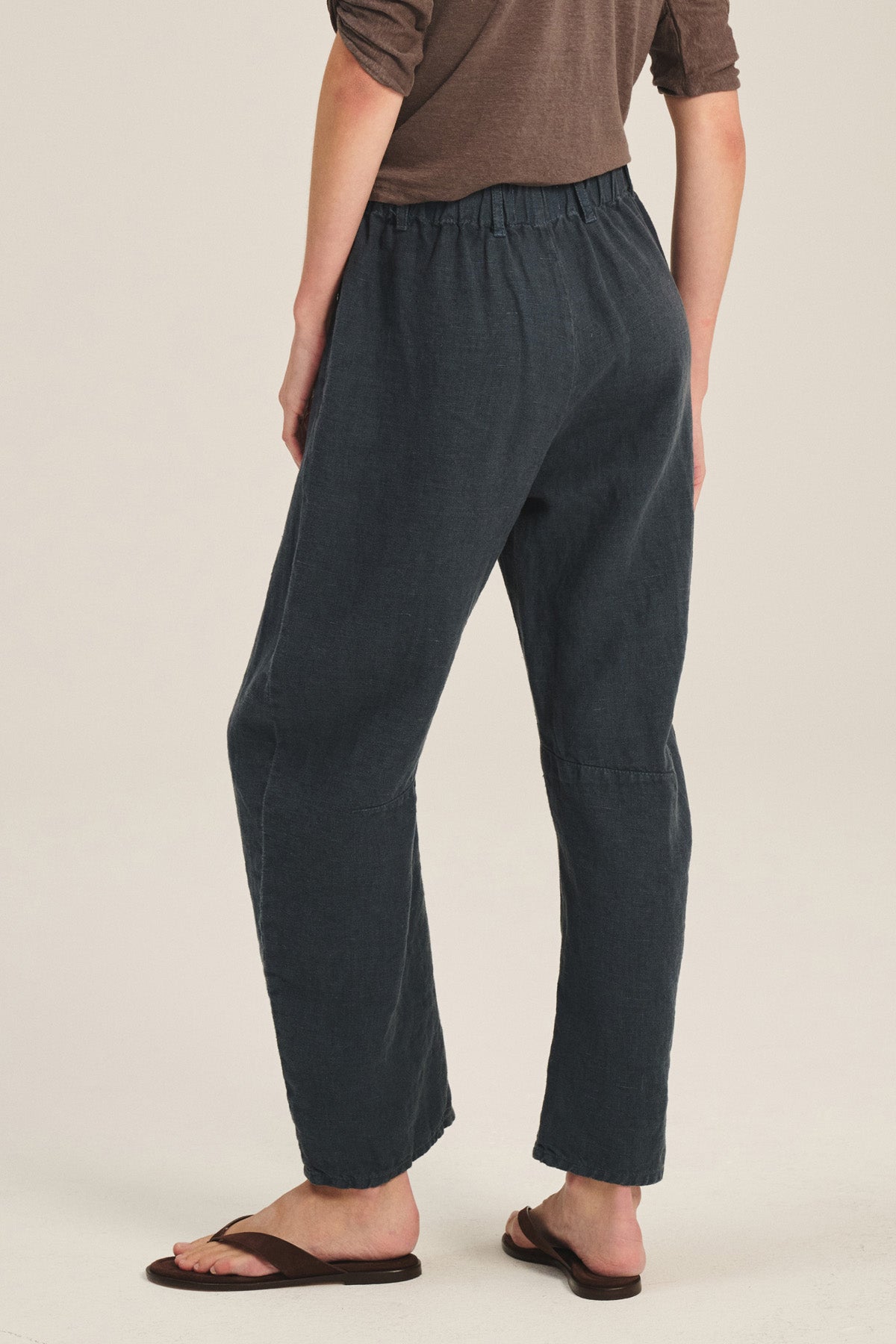 A person shown from the back wears a brown long-sleeve top, Velvet by Graham & Spencer TAYLOR HEAVY LINEN PANT featuring an elastic waistband and barrel leg, paired with brown sandals.