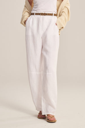Wearing Velvet by Graham & Spencer's TAYLOR HEAVY LINEN PANT—tailored with a curved leg—a person pairs it with a white tank, cream cardigan, brown belt, and sandals, standing hands in pockets.