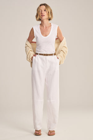 A woman stands against a plain background wearing Velvet by Graham & Spencer’s white TAYLOR HEAVY LINEN PANT, a white sleeveless top, beige cardigan draped on her arms, brown belt, and sandals.