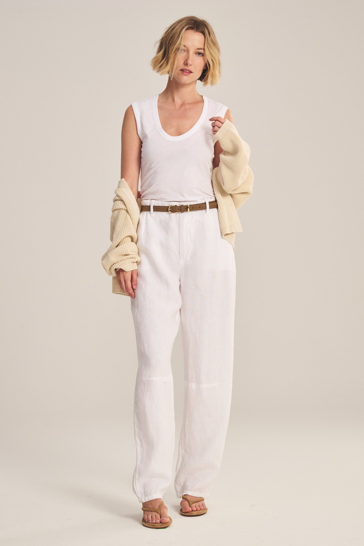 A woman stands in a studio wearing Velvet by Graham & Spencer's TAYLOR HEAVY LINEN PANT, paired with a white sleeveless top, brown belt, beige cardigan draped over her arms, and sandals.
