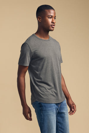 A man stands against a beige background wearing the HARRIS TEE by Velvet by Graham & Spencer—a gray triblend crew neck tee—paired with blue jeans, facing slightly to the right.