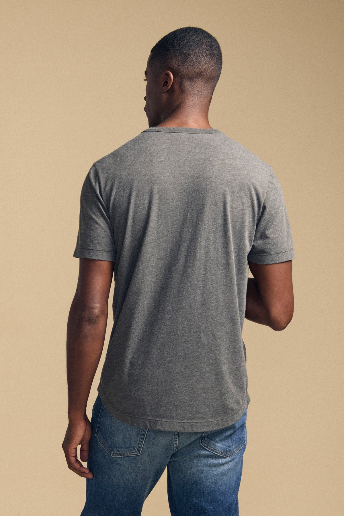 A man stands with his back to the camera, wearing the HARRIS TEE by Velvet by Graham & Spencer—a heathered gray short-sleeve t-shirt—paired with blue jeans against a beige background.