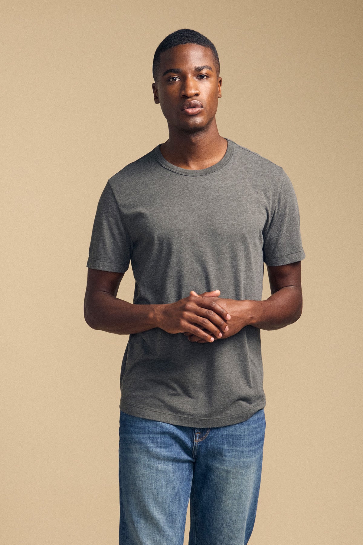 A man wearing the Velvet by Graham & Spencer HARRIS TEE in gray and blue jeans stands against a plain beige background, facing forward with his hands clasped.