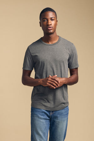 A man wearing the Velvet by Graham & Spencer HARRIS TEE in gray and blue jeans stands against a plain beige background, facing forward with his hands clasped.