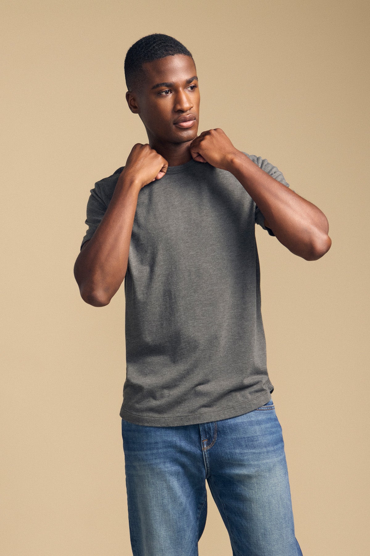 A person wears the HARRIS TEE in gray from Velvet by Graham & Spencer with blue jeans, standing against a beige background and holding the collar of the shirt with both hands.