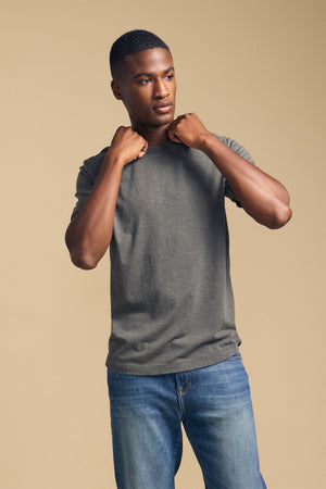 A person wears the HARRIS TEE in gray from Velvet by Graham & Spencer with blue jeans, standing against a beige background and holding the collar of the shirt with both hands.