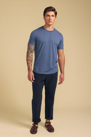 A man with a tattooed right arm wears the HARRIS TEE by Velvet by Graham & Spencer, paired with dark pants and brown sandals, standing against a plain beige background.