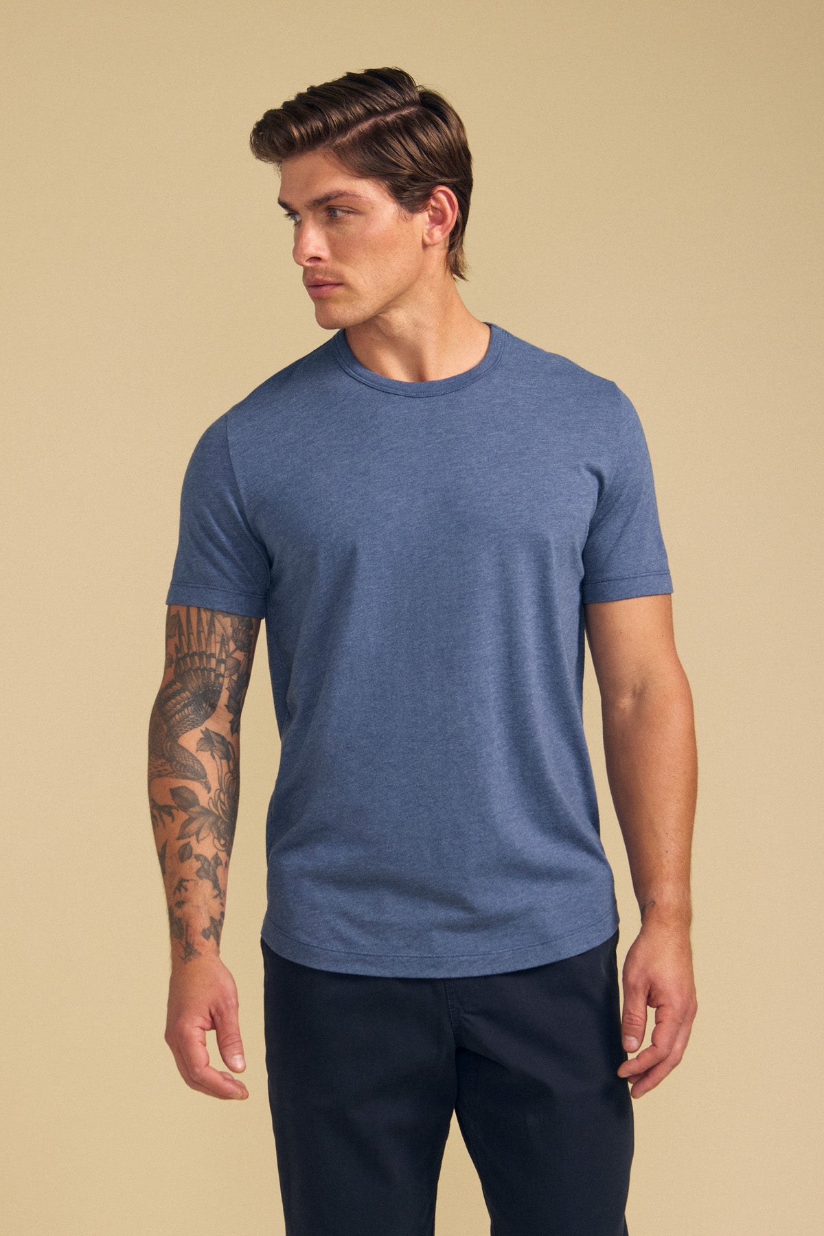 A man with a tattoo on his left arm wears the HARRIS TEE by Velvet by Graham & Spencer in blue and dark pants, standing against a beige background.
