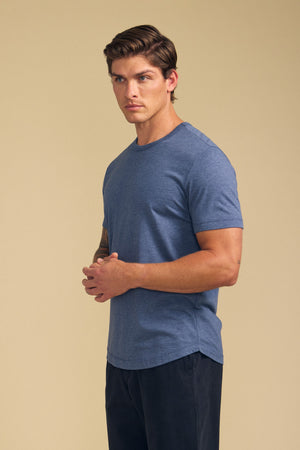 A man stands against a beige background, wearing the Velvet by Graham & Spencer HARRIS TEE in blue with dark pants, facing slightly to the side and loosely clasping his hands.