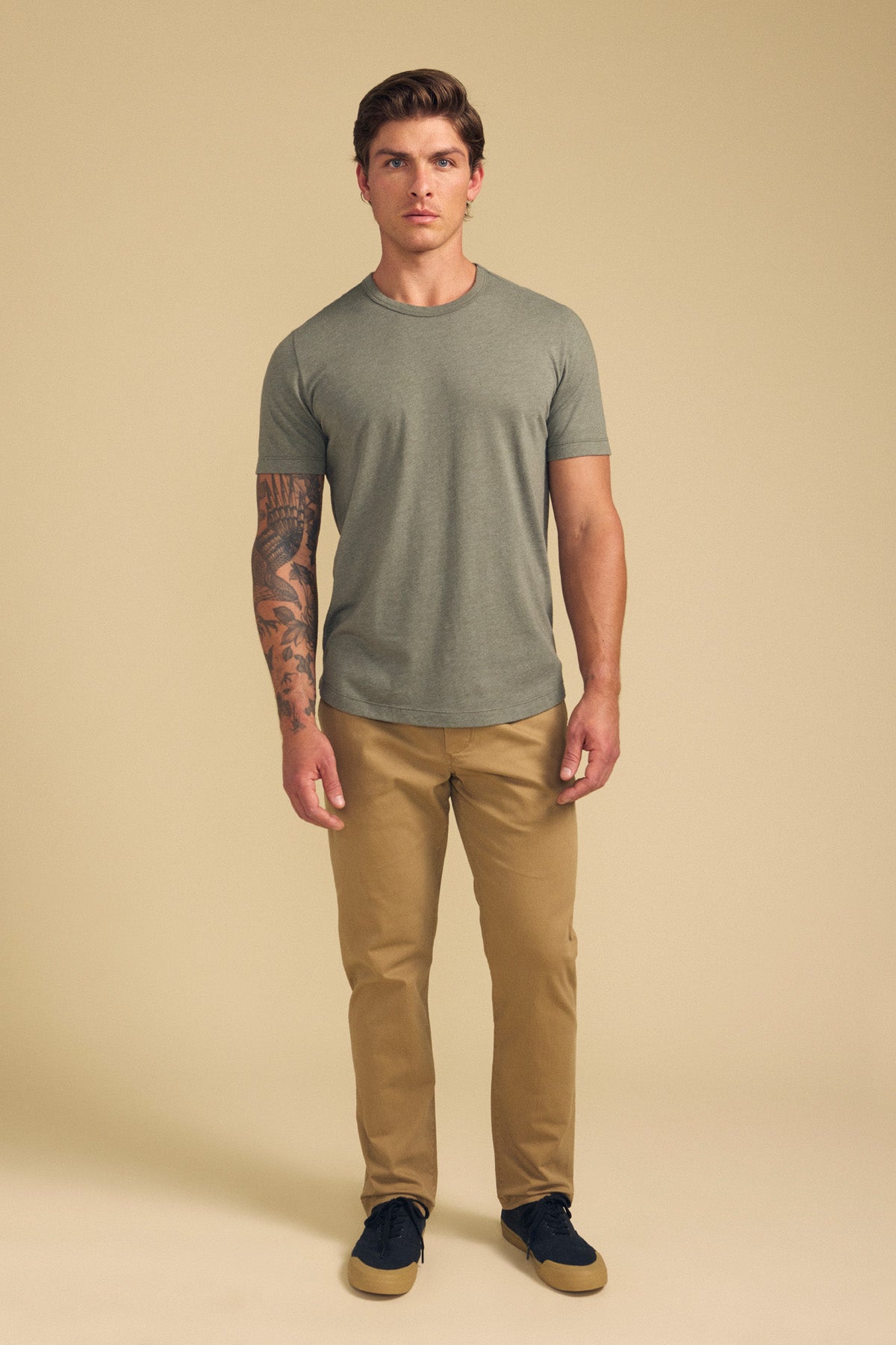 A man faces forward against a plain beige background, wearing the HARRIS TEE by Velvet by Graham & Spencer in gray, khaki pants, and black shoes; he has a tattoo sleeve on his left arm.-41702022774977