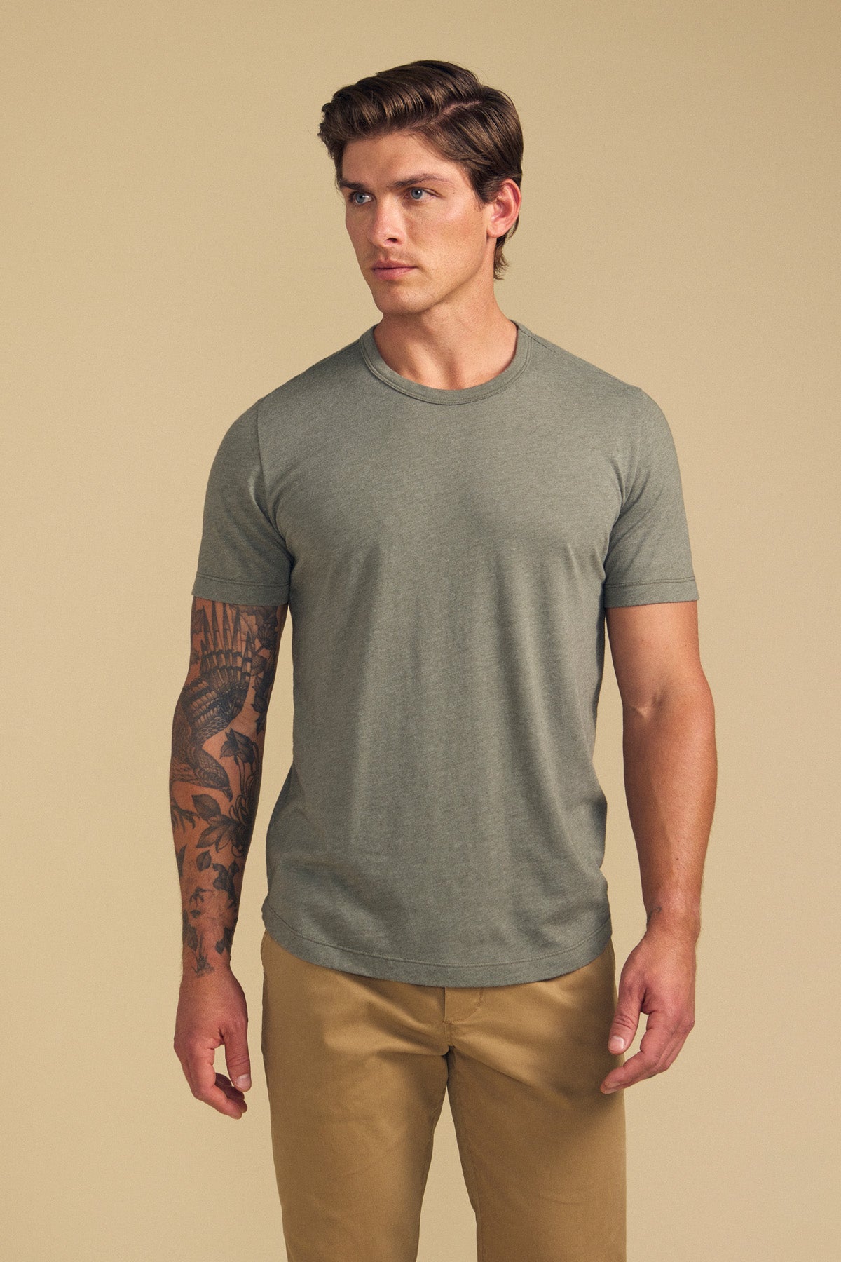 A man wearing the grey HARRIS TEE by Velvet by Graham & Spencer and khaki pants stands against a beige background, with a tattoo visible on his left arm.