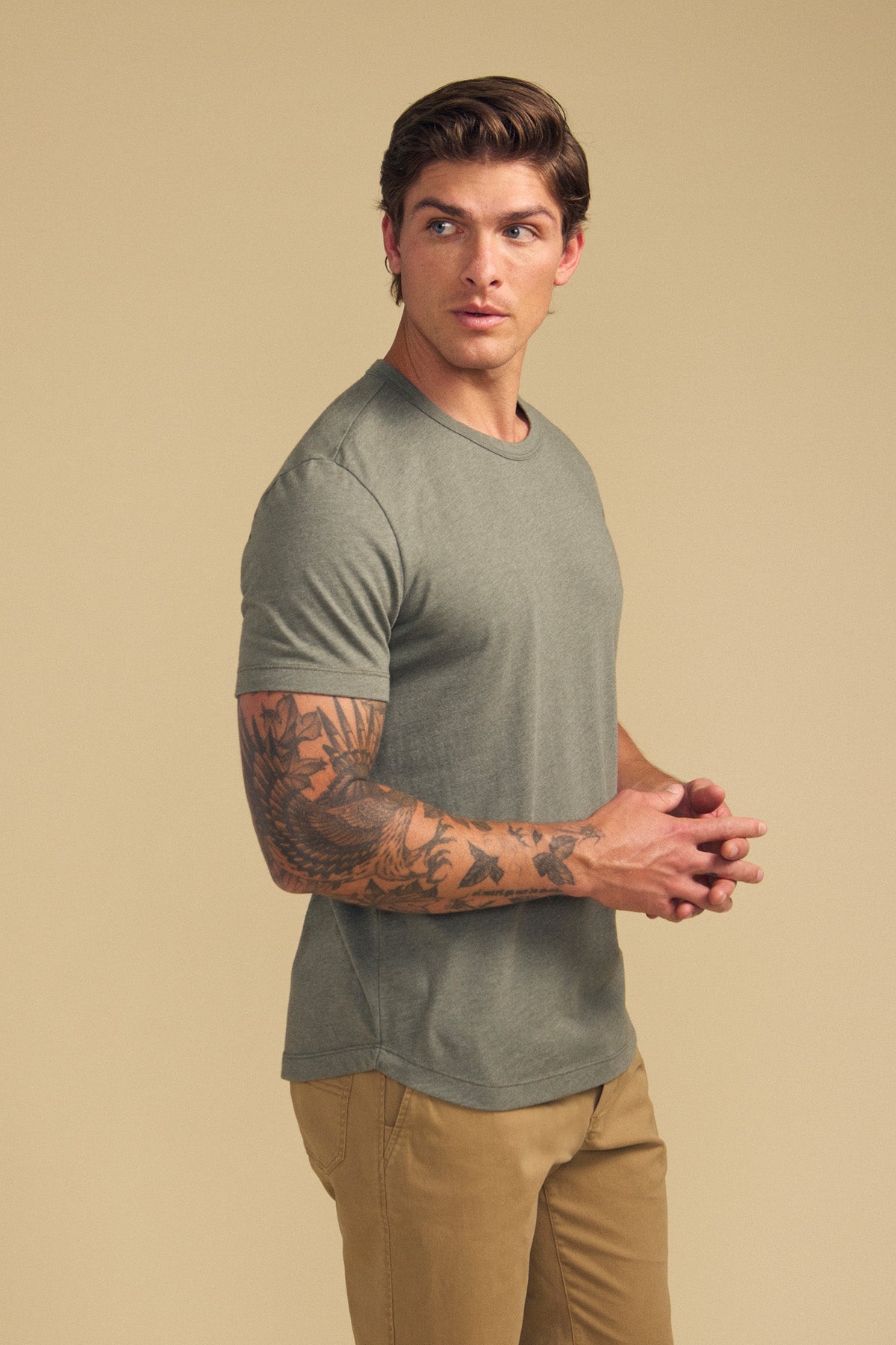 A man with tattooed arms models the HARRIS TEE by Velvet by Graham & Spencer in olive green and tan pants, standing against a beige background and looking to his left.