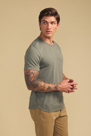 A man with tattooed arms models the HARRIS TEE by Velvet by Graham & Spencer in olive green and tan pants, standing against a beige background and looking to his left.