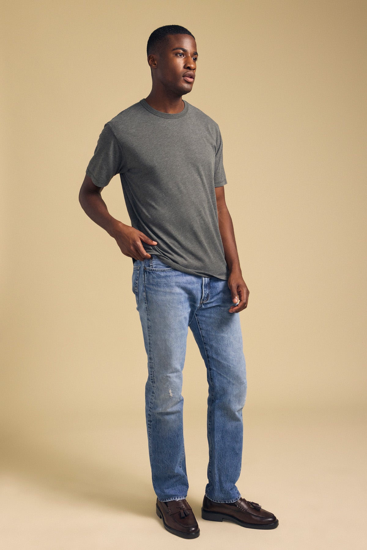 A man stands against a beige background wearing the Velvet by Graham & Spencer ZANDER TEE—a relaxed fit gray t-shirt with a classic crew neckline—paired with light blue jeans and brown shoes, one hand in his pocket.-42092518244545