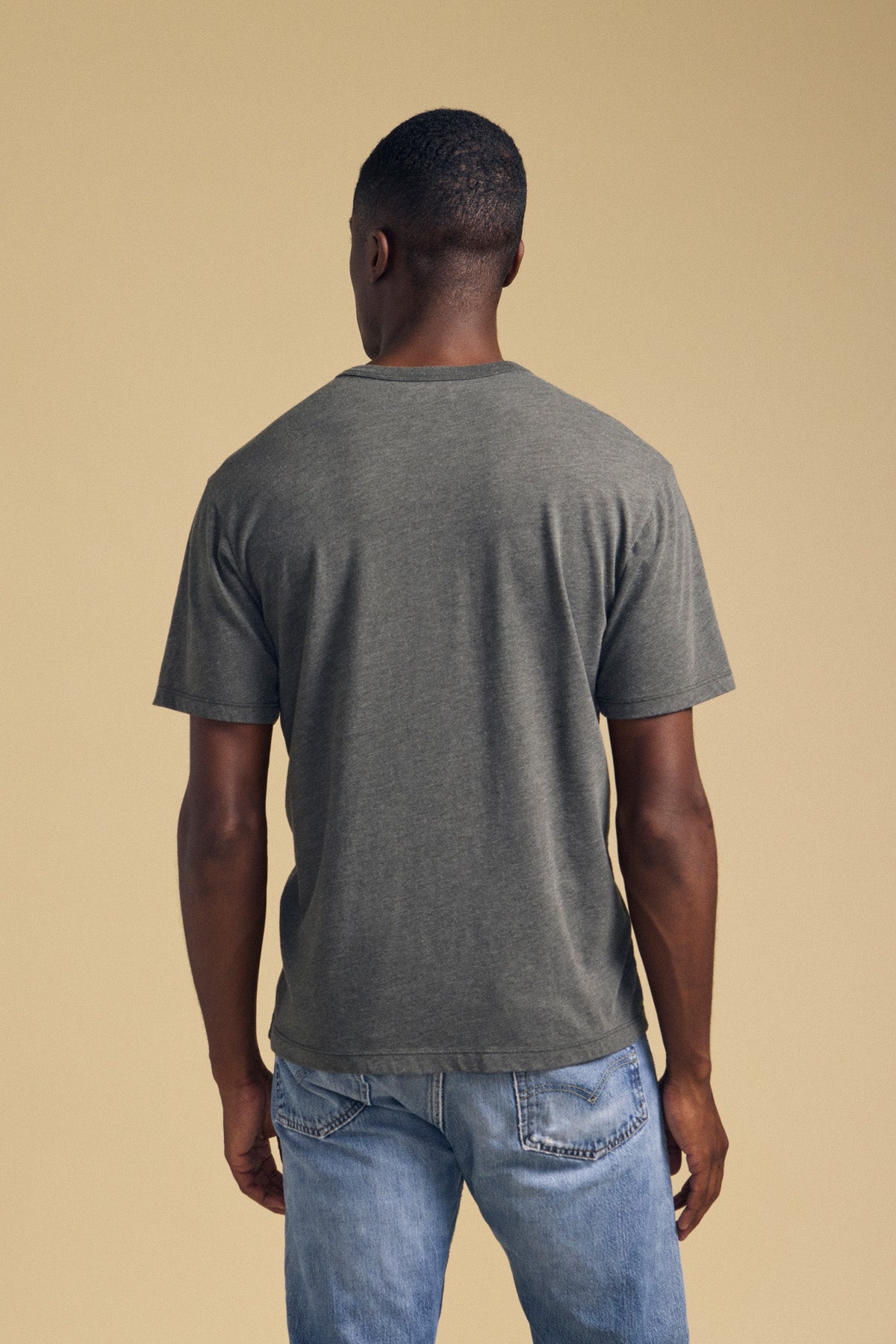 A person stands facing away from the camera, wearing the ZANDER TEE by Velvet by Graham & Spencer—a gray t-shirt with a classic crew neckline and relaxed fit, crafted from a soft cotton-modal blend for all-day comfort—paired with light blue jeans against a plain beige background.-42092518277313