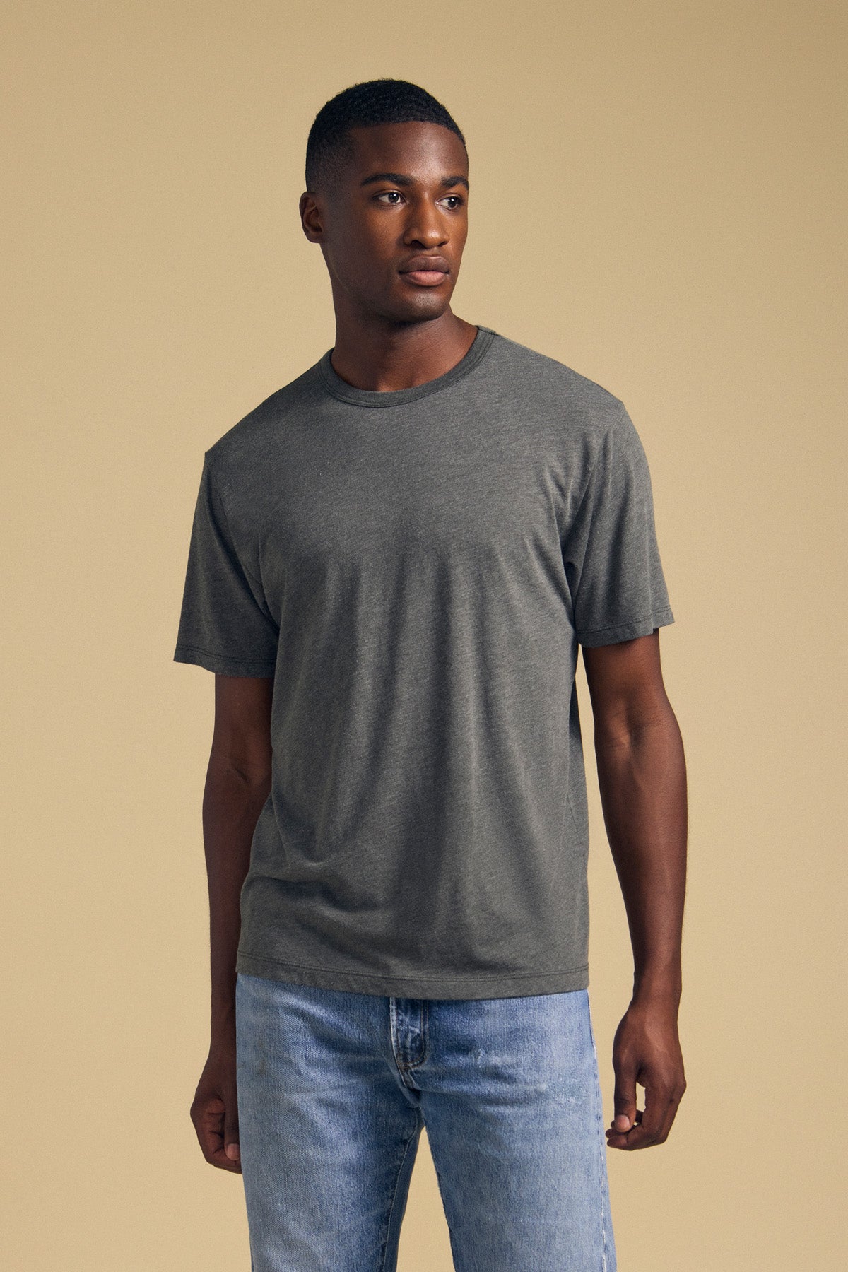 A man stands against a beige background wearing the ZANDER TEE by Velvet by Graham & Spencer—a plain gray cotton-modal blend t-shirt featuring a relaxed fit and crew neckline—paired with light blue jeans, facing slightly to the left.