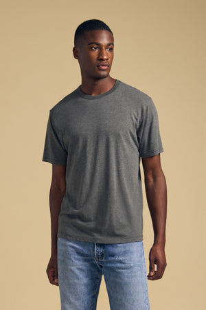 A man stands against a beige background wearing the ZANDER TEE by Velvet by Graham & Spencer—a plain gray cotton-modal blend t-shirt featuring a relaxed fit and crew neckline—paired with light blue jeans, facing slightly to the left.