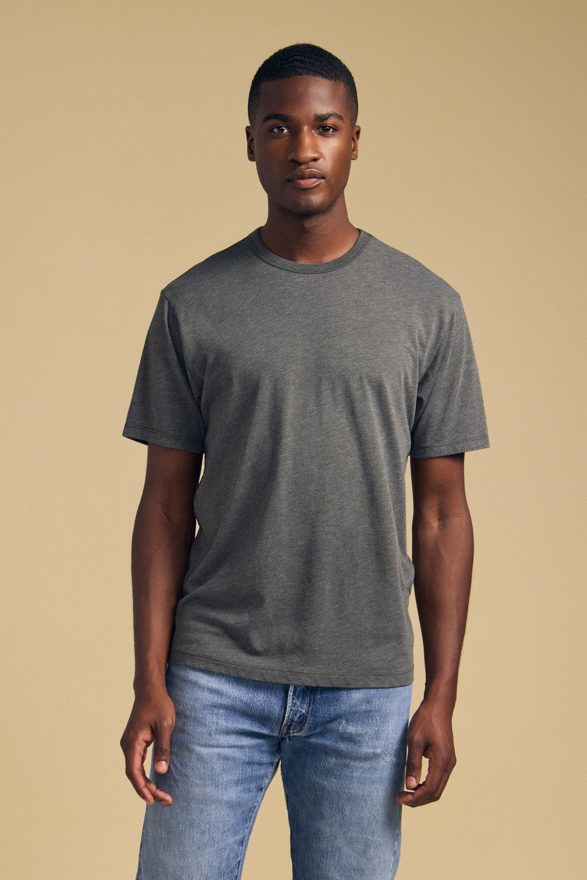 A person stands facing the camera wearing the Velvet by Graham & Spencer ZANDER TEE—a plain gray cotton-modal blend t-shirt with a classic crew neckline—and light blue jeans against a beige background.