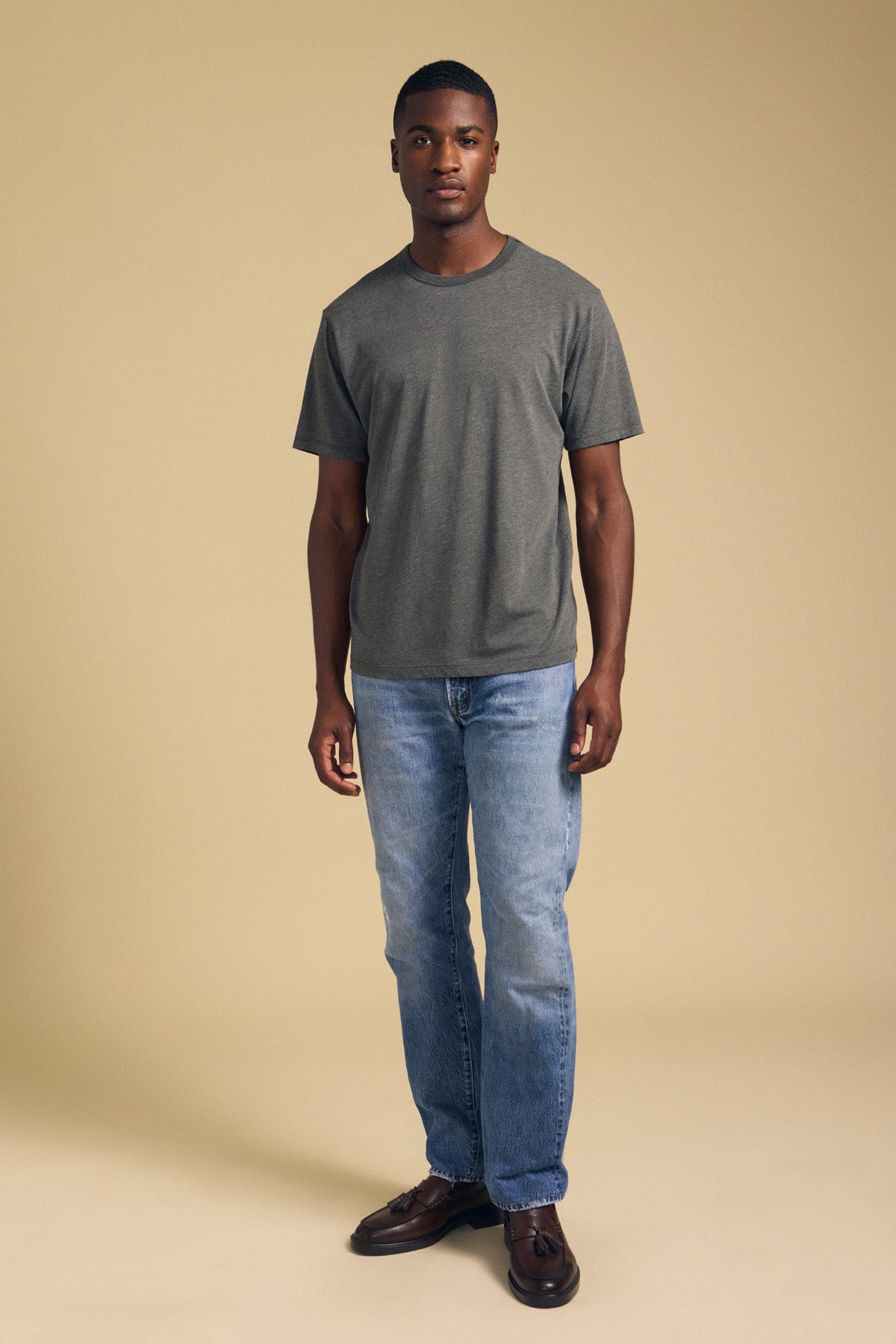 A man stands against a beige background, wearing the ZANDER TEE by Velvet by Graham & Spencer—a gray T-shirt featuring a classic crew neckline and relaxed fit—paired with light blue jeans and brown shoes. Crafted from a soft cotton-modal blend, the tee offers all-day comfort.-42092518080705