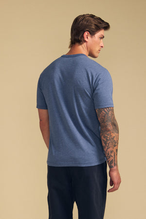 Man with his back to the camera wears the Velvet by Graham & Spencer ZANDER TEE—blue, relaxed fit, short-sleeve in a soft cotton-modal blend—with dark pants and a visible right arm tattoo, set against a beige background. Perfect for everyday wear.