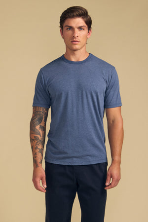 A man wears the Velvet by Graham & Spencer ZANDER TEE, a blue relaxed-fit short-sleeve shirt, with dark pants. He stands facing forward against a beige backdrop, his left arm tattoo visible—effortless everyday style.