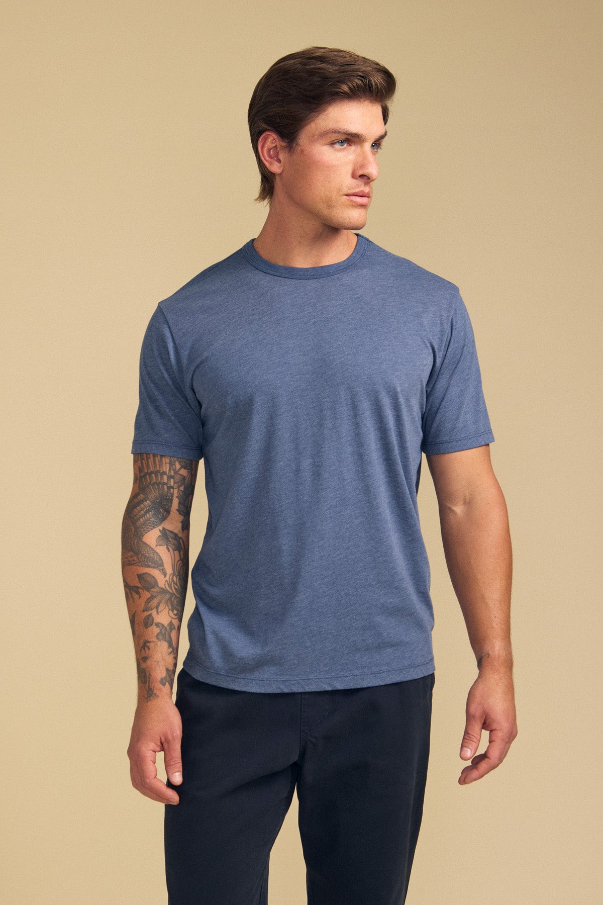 A man with a tattoo on his left arm wears the Velvet by Graham & Spencer ZANDER TEE in blue, paired with dark pants, standing against a plain beige background.-41702129959105