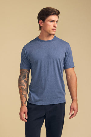 A man with a tattoo on his left arm wears the Velvet by Graham & Spencer ZANDER TEE in blue, paired with dark pants, standing against a plain beige background.